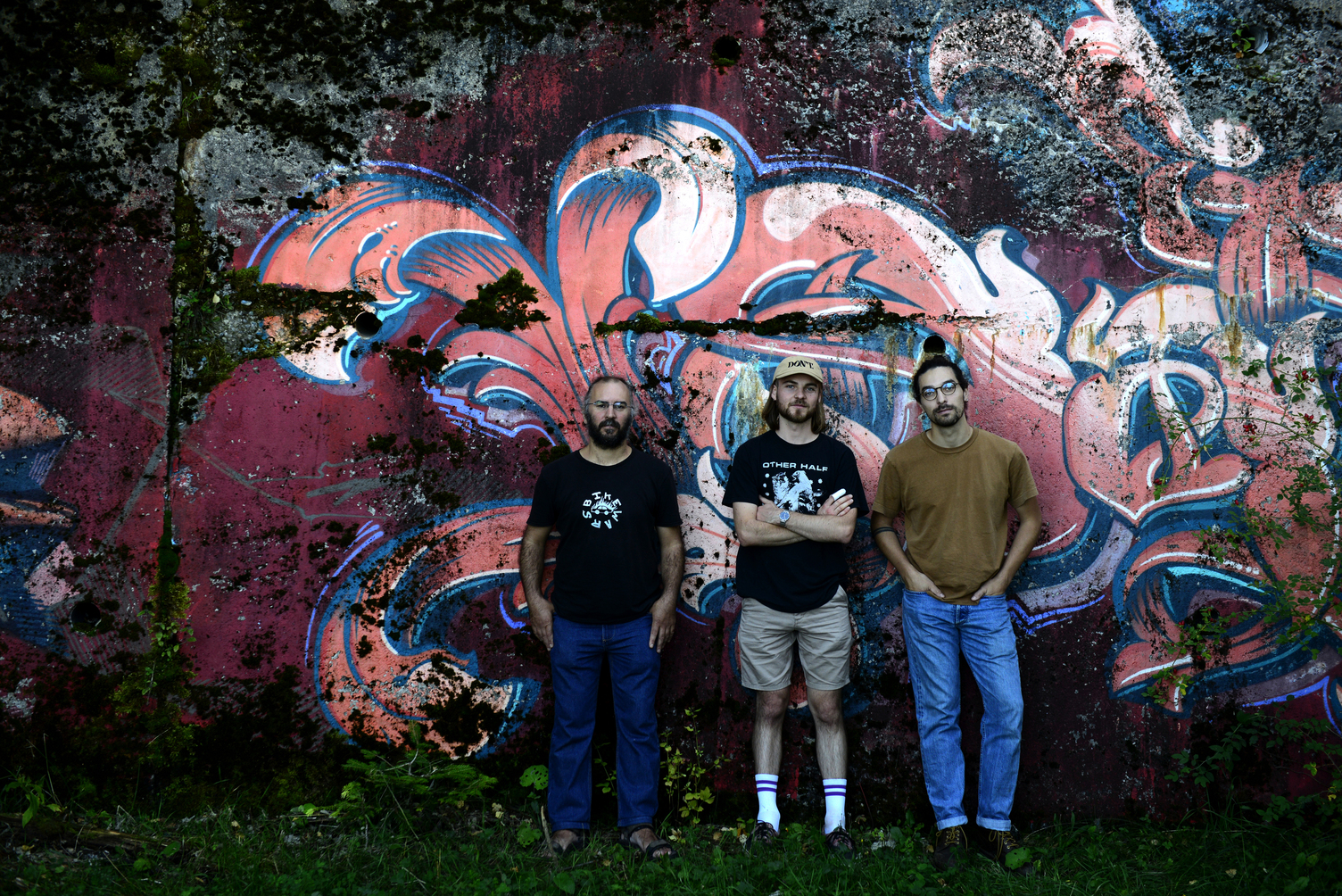 Les membres de Moyō posent devant un mur de béton recouvert d’une fresque graffiti rose et bleue.